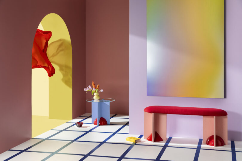 A vibrant interior setup featuring a red bench, white square flooring, a small table with a vase, abstract art on the walls, and a floating red scarf.
