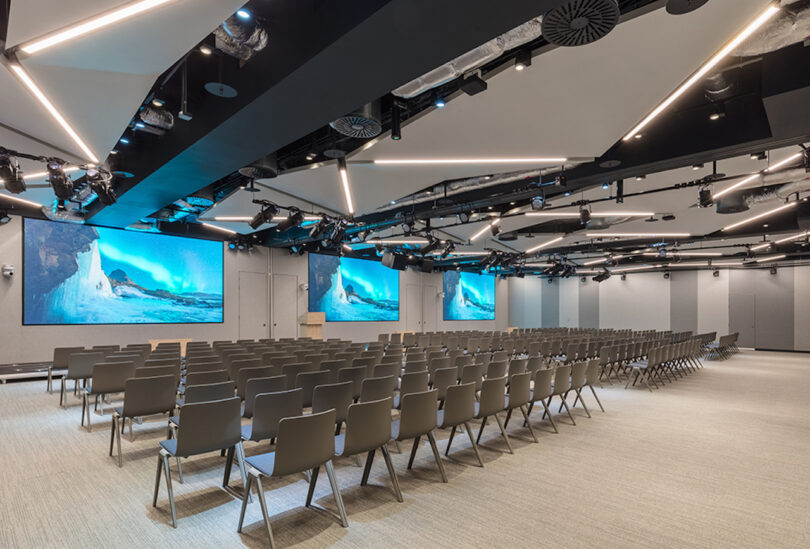 A modern conference room with rows of empty chairs facing two large screens displaying a nature scene. The ceiling features geometric lighting fixtures and various audio-visual equipment