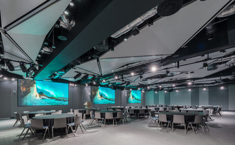 A spacious, modern conference room with multiple round tables, a podium, and large screens displaying an image of a glacier. The ceiling features exposed pipes and high-tech lighting