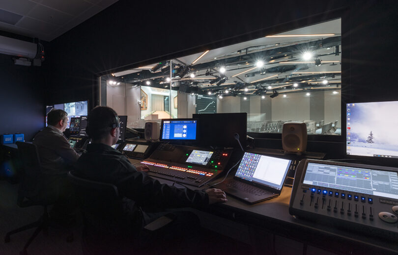 Two technicians work at a control panel with multiple screens in a dimly lit studio, overseeing a large, well-lit auditorium