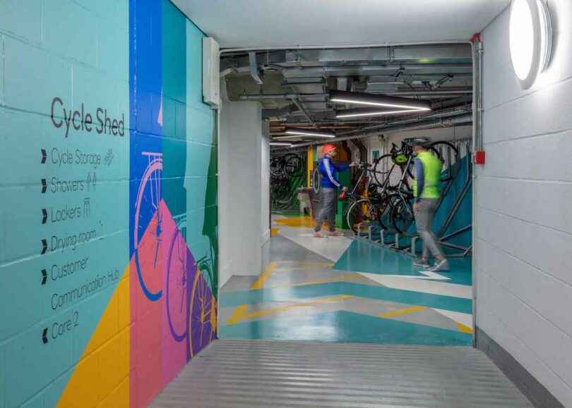 A brightly colored cycle shed with bikes stored on racks. The wall lists available facilities: cycle storage, showers, lockers, dining room, customer communication hub, and care 2. Two people are present