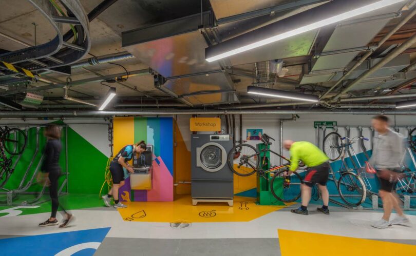 People in an indoor bike workshop performing maintenance tasks on their bikes. The space is equipped with tools, workstations, and bike racks, and features colorful wall art