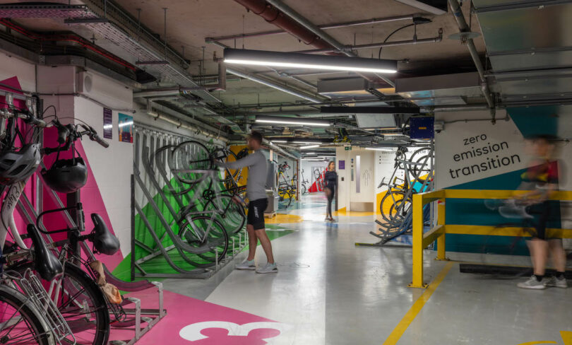 People park and retrieve bicycles in a modern, indoor bike storage area with painted walls and signs promoting zero emission transition