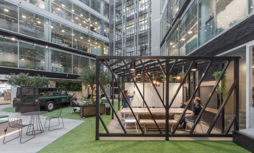 An open courtyard with glass walls features a wooden gazebo, office table with chairs, potted plants, a vintage vehicle, and people seated on benches