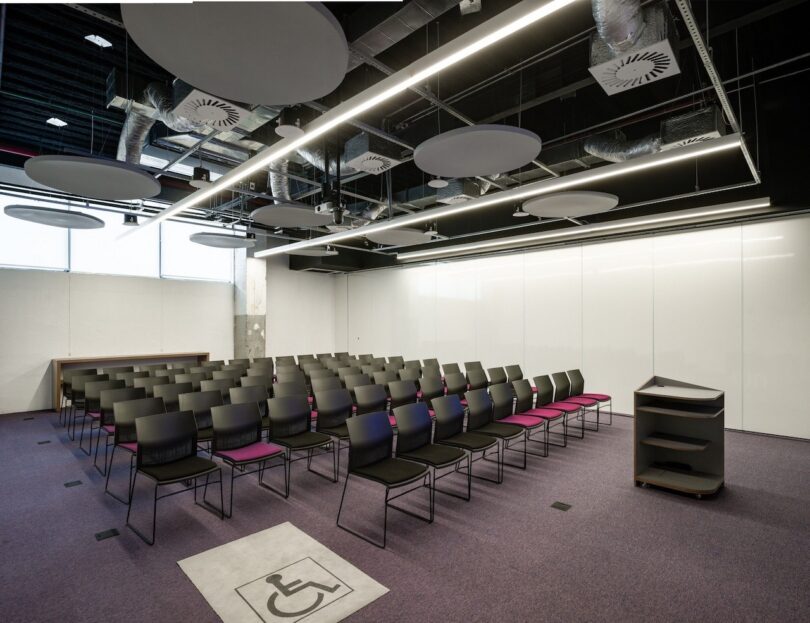 Modern conference room with rows of black chairs, some with pink accents, a wheelchair accessible space, and circular lighting fixtures on the ceiling