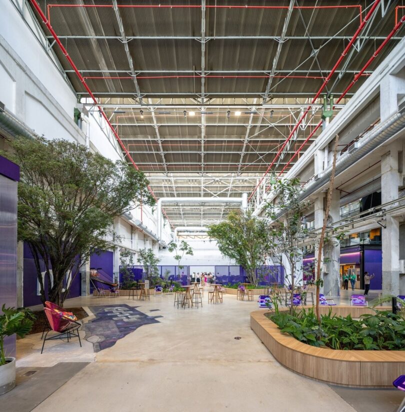 Interior of a modern open-air office with purple accents, featuring green plants, seating areas, and a high industrial ceiling