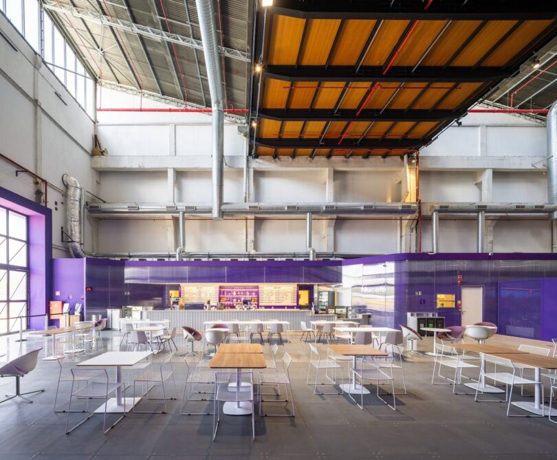 Spacious industrial-style cafeteria with purple accents, exposed ductwork on ceiling, and various seating arrangements