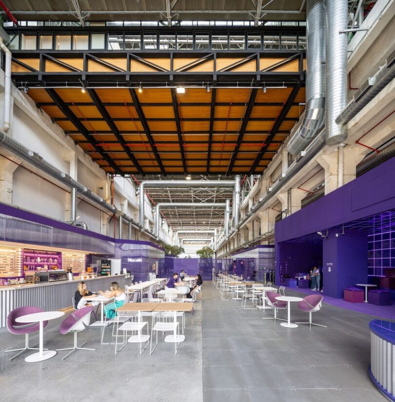 Modern indoor cafe with vibrant purple accents, people dining at tables, and exposed ceiling pipes