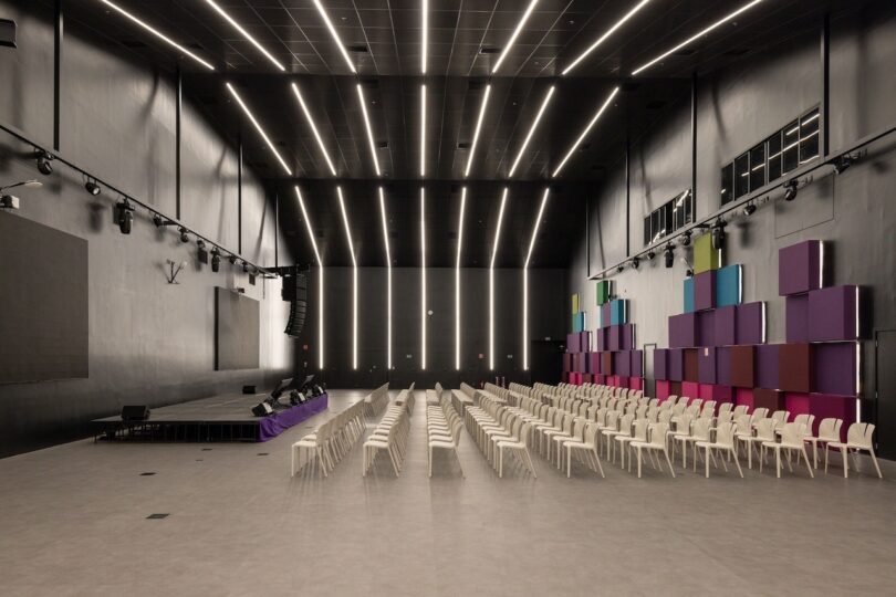 Modern auditorium with rows of white chairs facing a stage, surrounded by black walls and colorful acoustic panels under a ceiling with linear lights