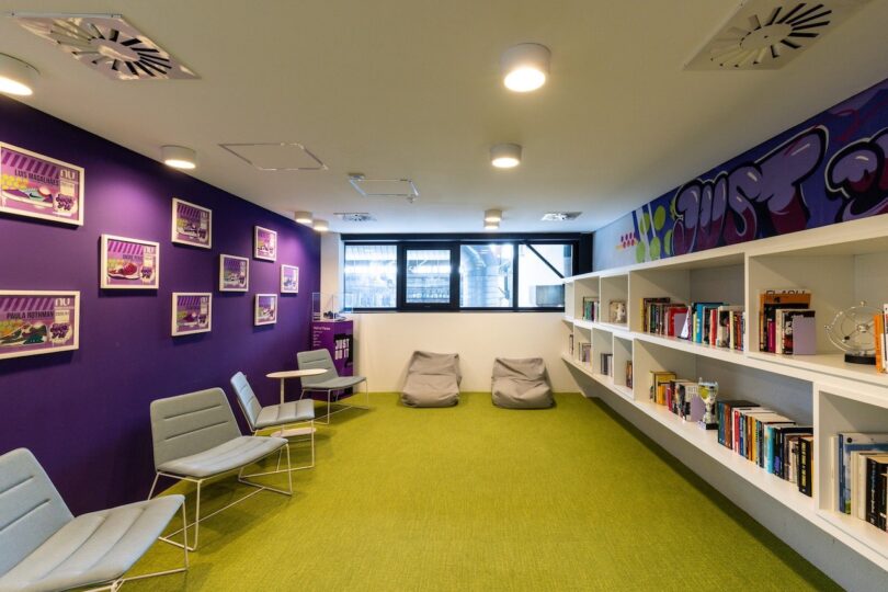 Modern library room with vibrant purple walls, white shelves filled with books, green carpet, and large window, furnished with bean bags and metal chairs