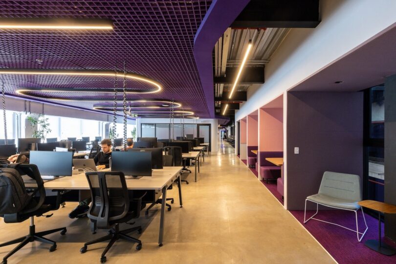 Modern office space featuring desks with computers, ergonomic chairs, purple walls, and circular hanging lights