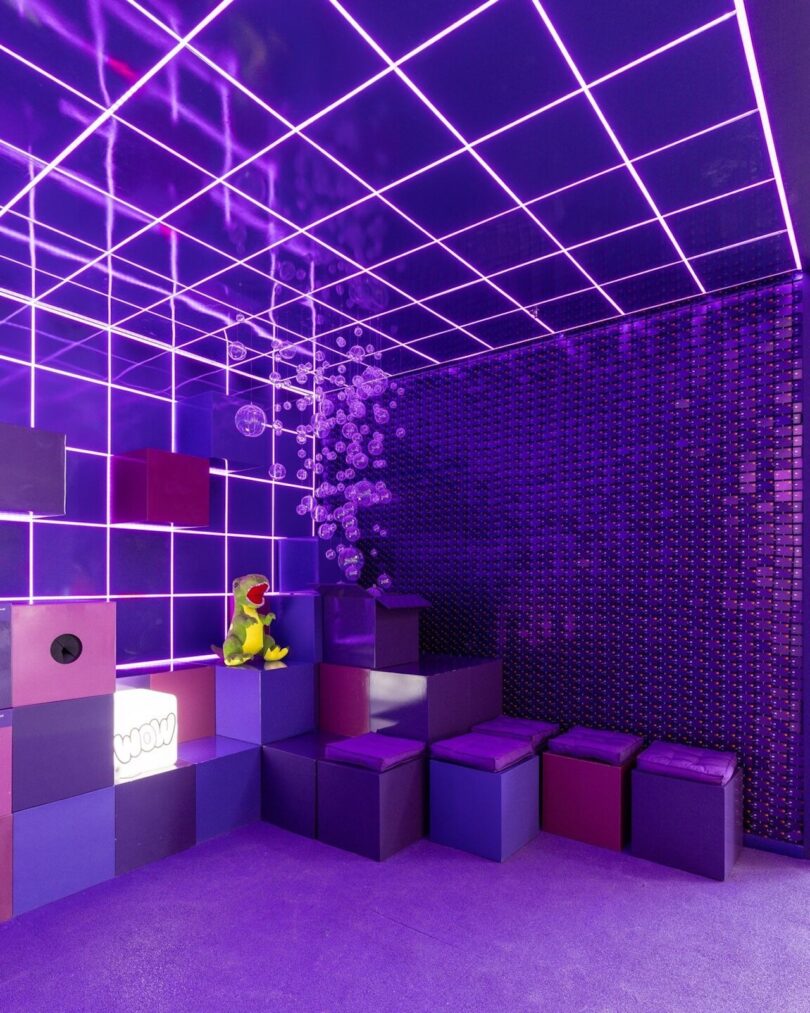 A bright purple room featuring grid light patterns on the ceiling and walls, decorated with a green plush toy frog, geometric blocks, and hanging clear bubbles