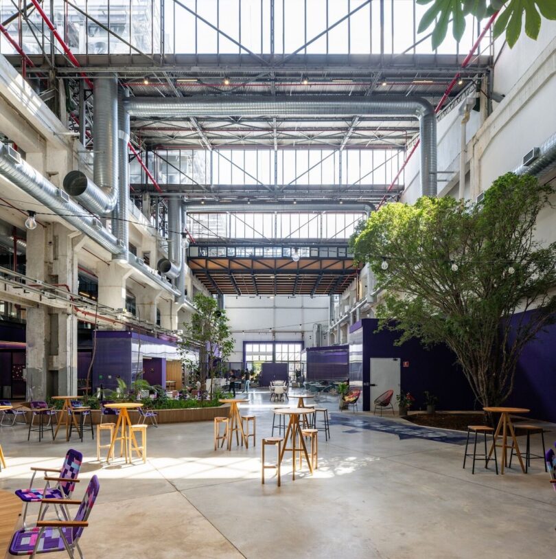 Modern industrial-style lobby with exposed ceiling beams and ductwork, featuring a variety of seating and large, leafy plants, bathed in natural light from skylights