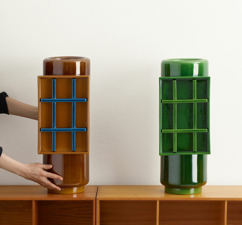Two cylindrical vases, one brown and one green, with gridded inserts on wooden shelves. A hand is adjusting the brown vase on the left