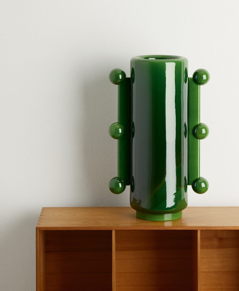 A glossy green ceramic vase with round protrusions on both sides, placed on a light wooden shelf with cubbyholes against a plain white wall