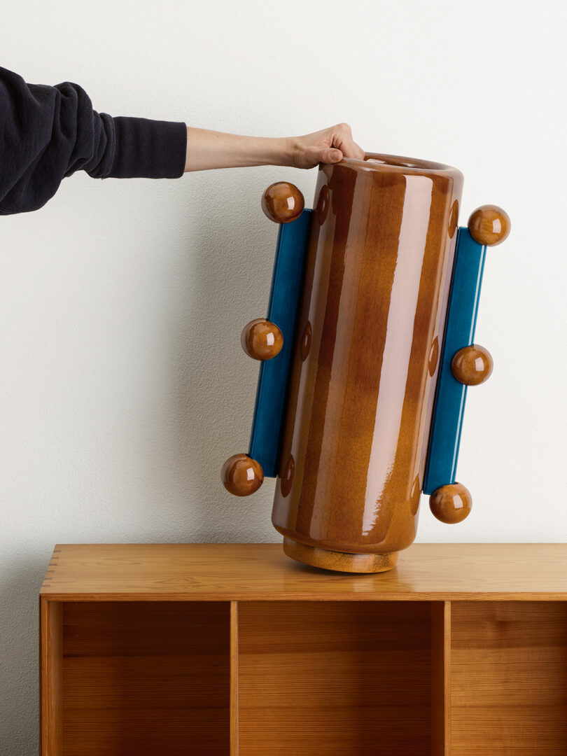 A person holds a large, cylindrical vase with blue handles, placing it on a wooden shelf unit.