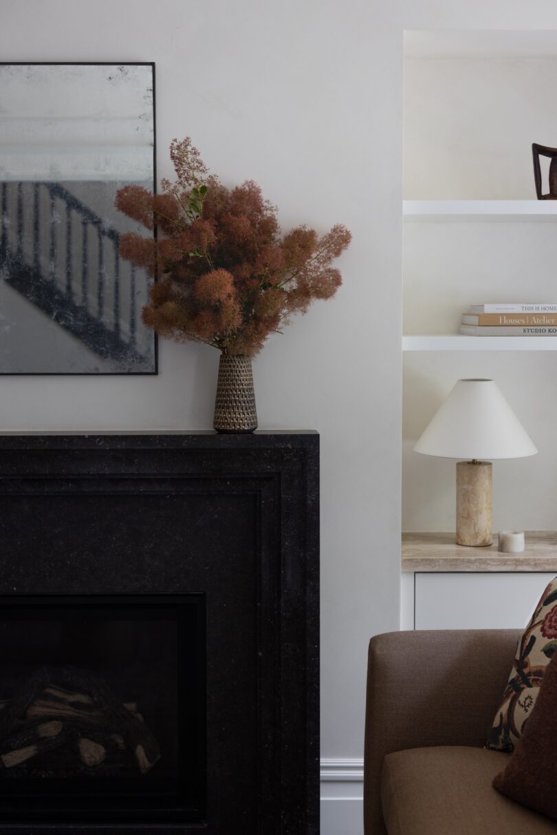 A close-up of a living room corner featuring a black fireplace with a textured vase of dried flowers on the mantel.