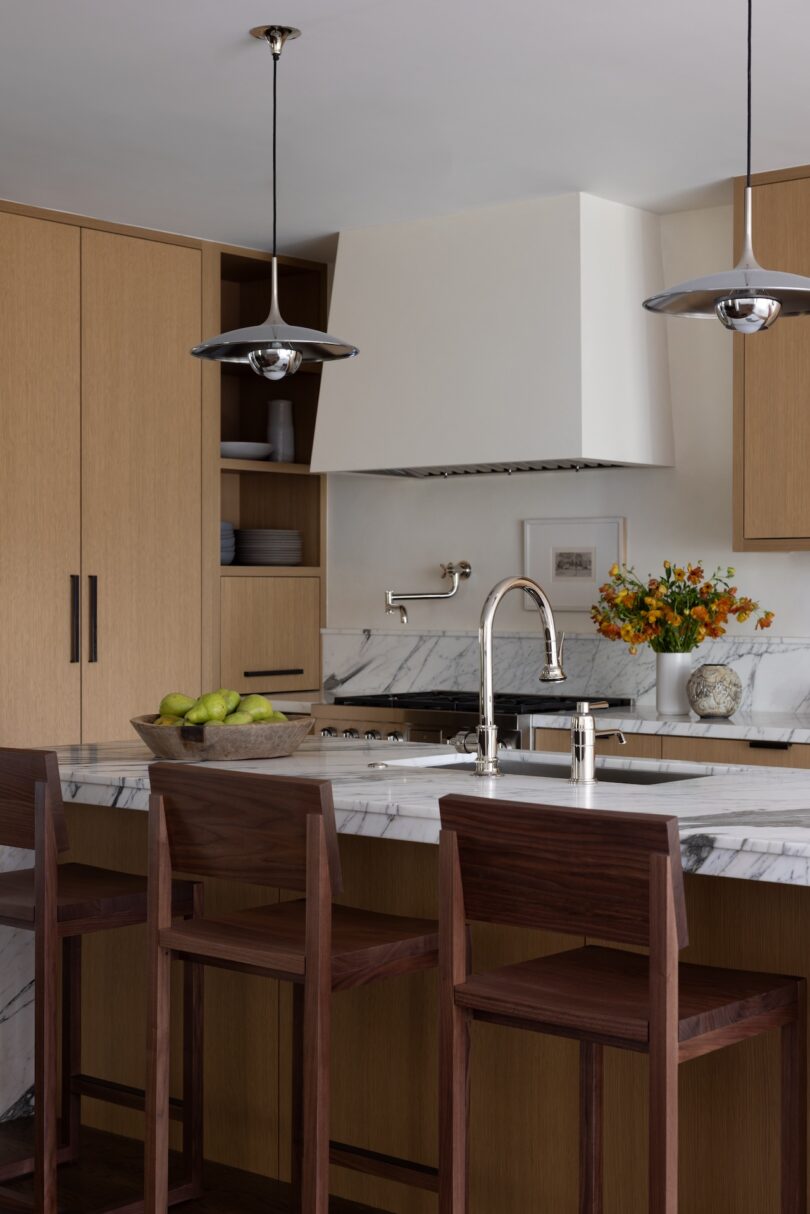 A modern kitchen with light wood cabinetry and a large marble island. Three wooden bar stools with dark wood backs and seats line the island.