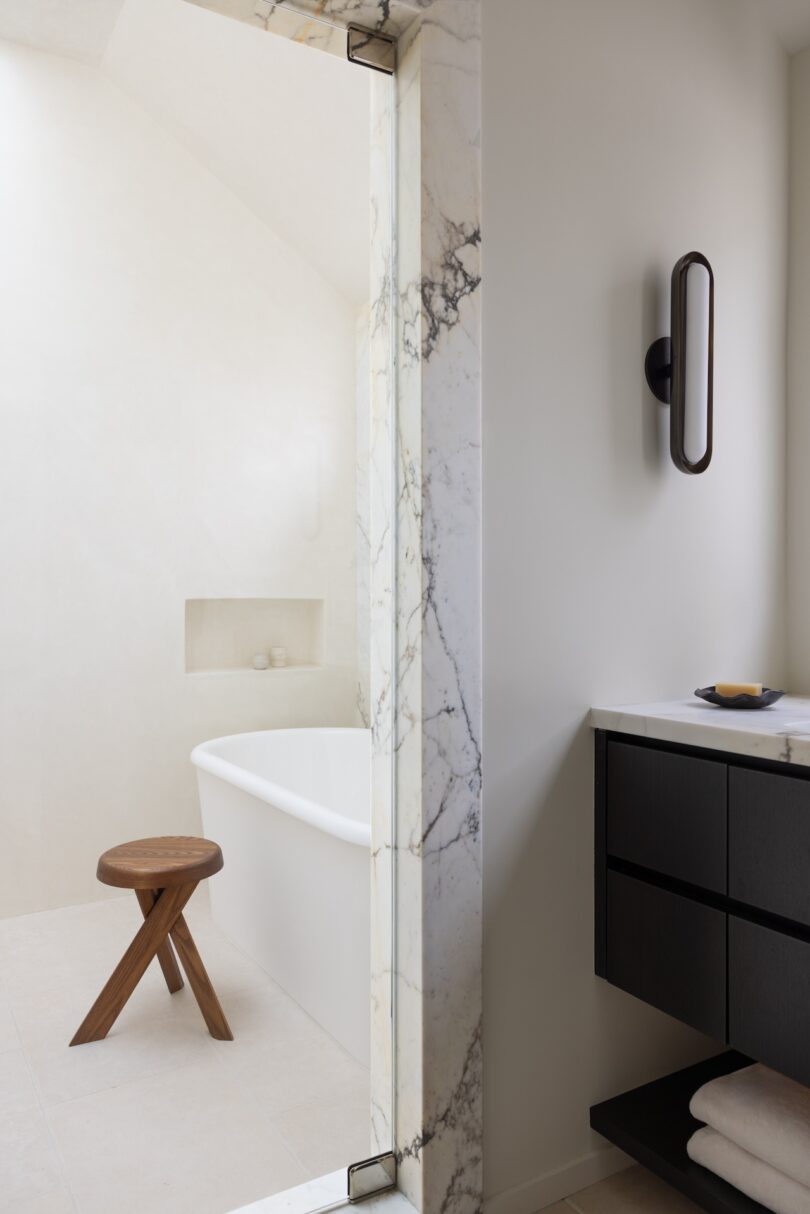 A minimalist bathroom featuring a white freestanding bathtub, a marble-framed glass shower door, and a small wooden stool.