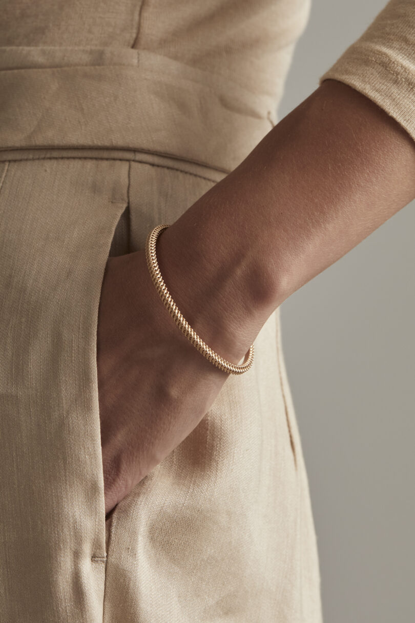 Close-up of a person's arm wearing a gold bracelet, with a hand on the hip, dressed in a beige linen outfit