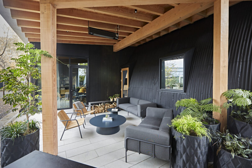 Modern outdoor patio with gray furniture and lush green plants, featuring a wooden ceiling and black walls