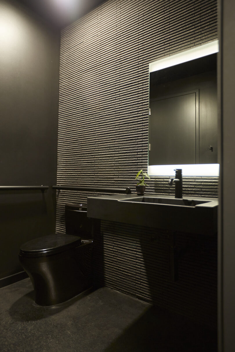 Modern bathroom with black fixtures, including a toilet and sink, lit by a backlit mirror on a textured dark wall