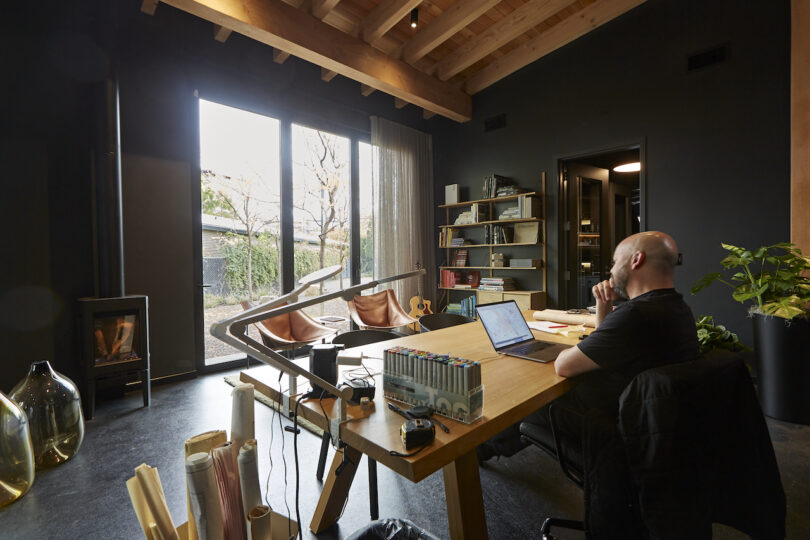 A man sitting at a wooden desk in a modern home office, with a laptop, books, and personal items. the room has dark walls, wooden ceilings, and a fireplace