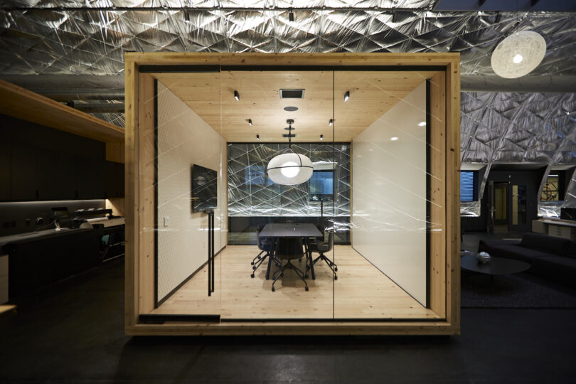 Modern office meeting room enclosed in a glass and wooden frame, featuring a large table, chairs, and unique overhead lighting, surrounded by an open workspace