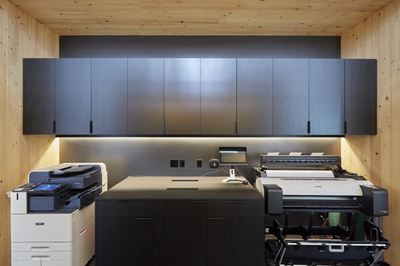 Modern office print station with a white multifunction printer, a large format canon printer, under soft lighting and black storage cabinets, surrounded by wooden walls