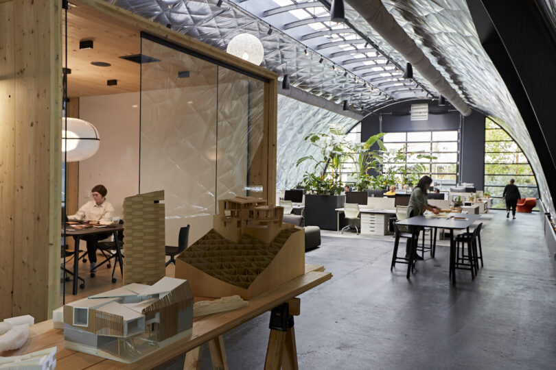 Modern architectural office with employees working, featuring large models and plants, enclosed by glass partitions under an arched ceiling.