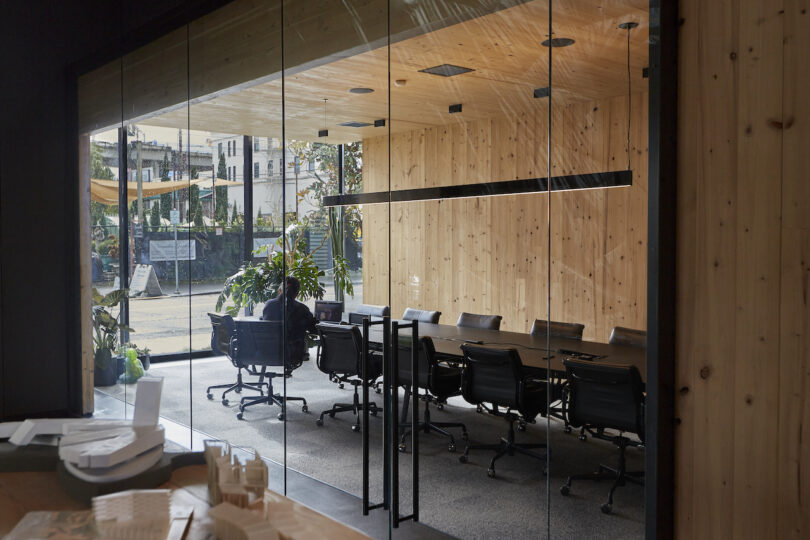 Modern office conference room with glass walls, wooden panels, and a long table with chairs, overlooking an outdoor view