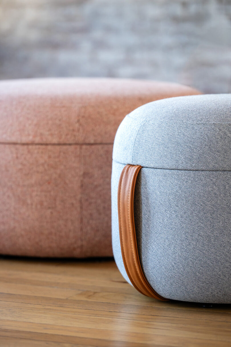 Two round fabric ottomans, one in light pink and the other in grey with a brown leather handle, placed on a wooden floor