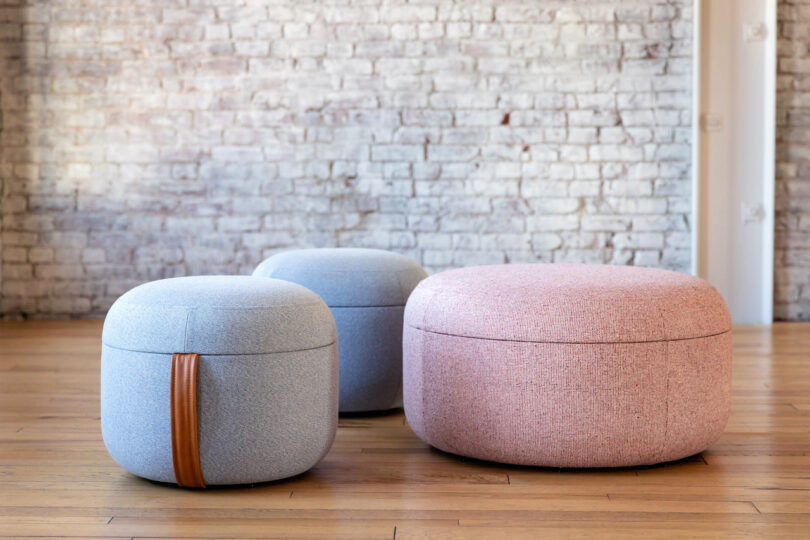 Three round ottomans, two small in light blue and one large in light pink, are placed on a wooden floor in front of a white brick wall