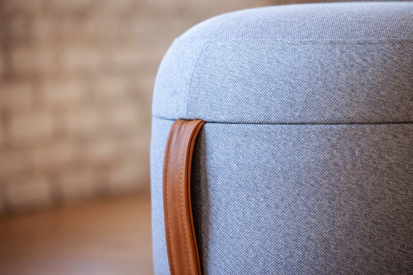 Close-up of a light grey fabric ottoman with a brown leather handle. The background is out of focus