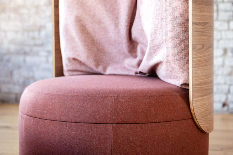A close-up view of a modern, wooden-framed chair with light red upholstery and a cushioned backrest, in front of a textured brick wall