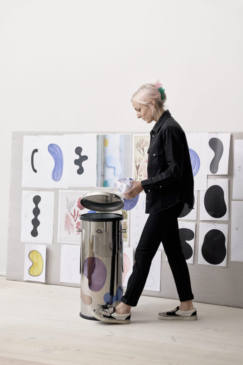 woman opening a steel trash can with magnetic shapes attached to it with her foot in front of a wall with sketches taped up