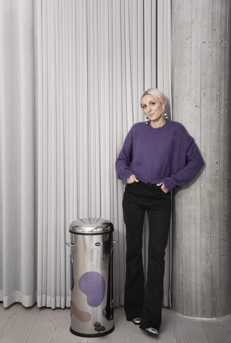 Woman in purple sweater and black jeans standing next to a metal trash can 