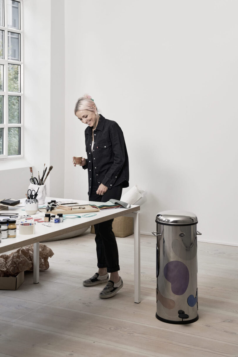 A woman in a black shirt stands by a desk, examining sketches in a bright, minimalist studio with art supplies scattered around next to a steel trash bin