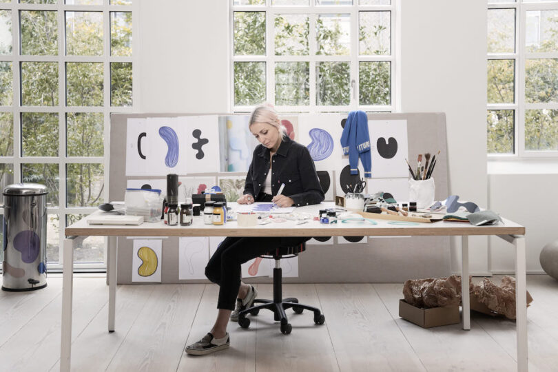 A woman works at a desk in a bright art studio with large windows, surrounded by colorful sketches and various art supplies