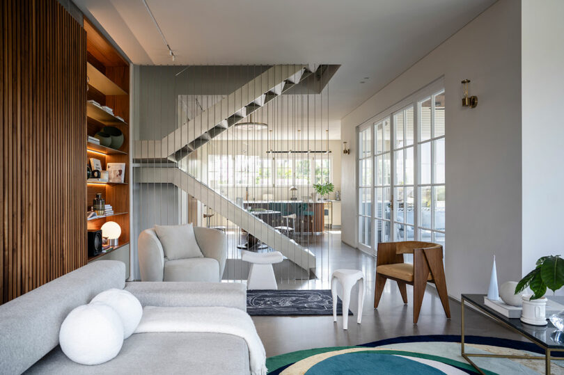 A modern living room features a gray sofa, armchair, wooden accent chair, and a staircase with vertical railings. Large windows on the right let in natural light, highlighting the contemporary decor.