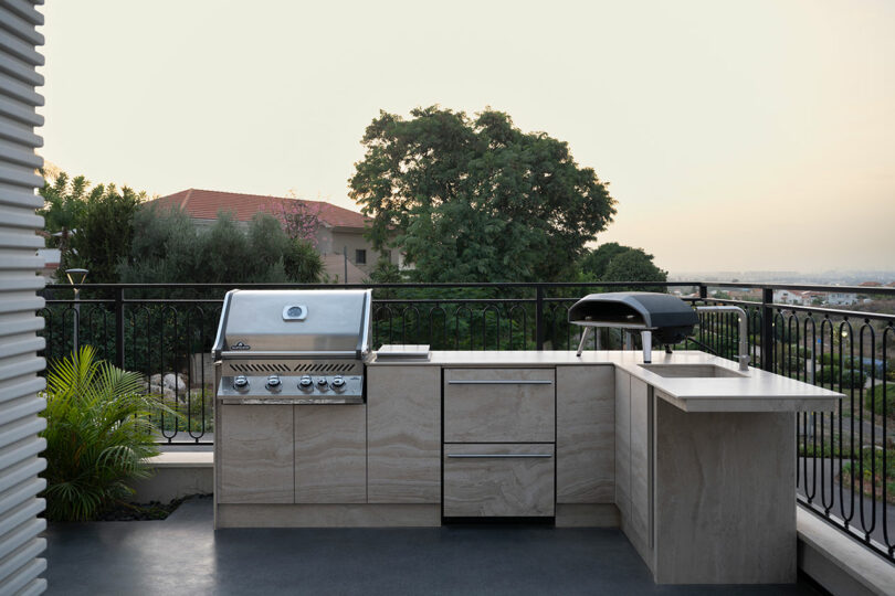 Outdoor kitchen with a grill, countertop, drawers, and a sink overlooking greenery and houses in the distance.