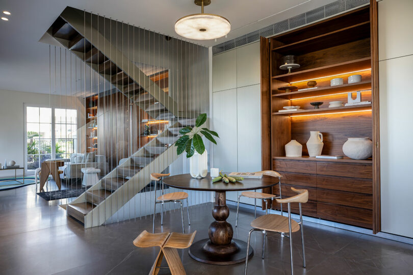Modern kitchen with wooden dining table, chairs, and an open shelving unit displaying ceramics. A sleek staircase with vertical cables leads to the upper level. A large window allows natural light in.