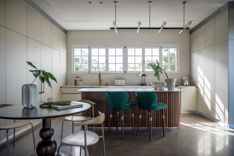 A modern kitchen with a large island featuring green bar stools, a wooden table with chairs, and large windows providing natural light. Decor includes plants and various kitchen accessories.
