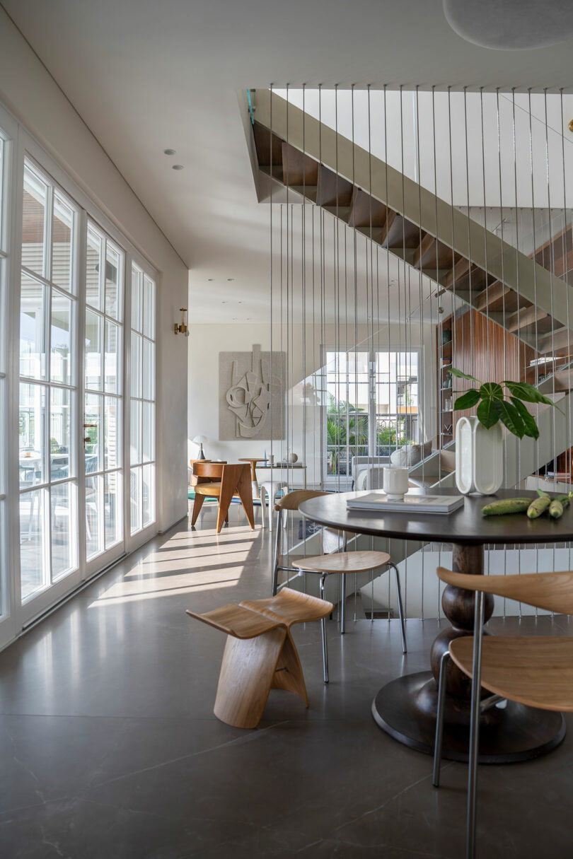 Modern dining area with floor-to-ceiling windows, a table with wooden chairs, minimalist decor, and a staircase with thin vertical railings leading to an upper floor. The room is well-lit with natural light.
