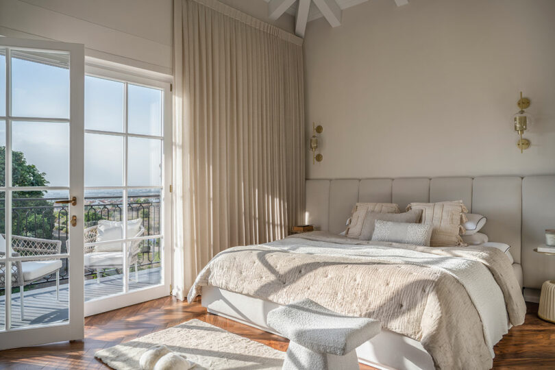 A sunlit bedroom with a large bed, light beige bedding, and a cushioned headboard. A glass door opens to a balcony with a view of greenery.
