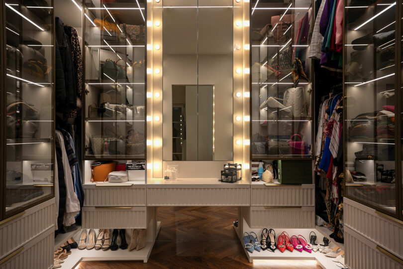 A modern walk-in closet showcasing illuminated glass cabinets filled with handbags and other accessories, a central vanity with lighted mirror, and neatly arranged shoes below.