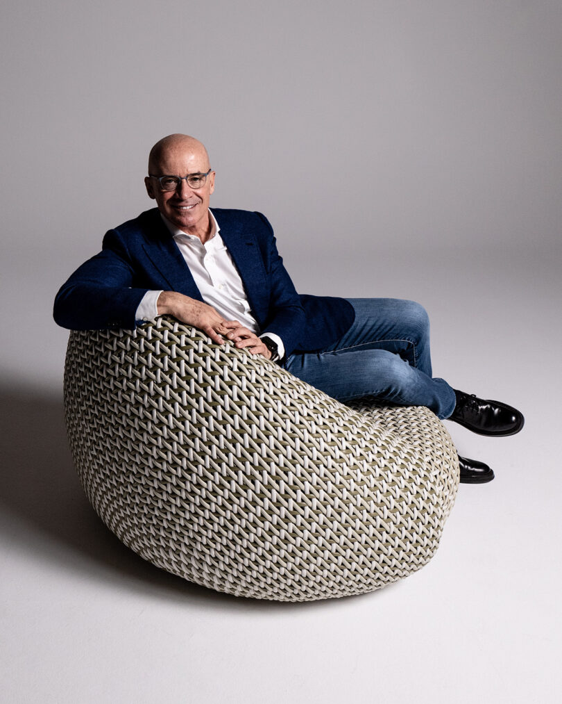 A bald man in a blue blazer, white shirt, and jeans sits in a textured, woven chair with one arm resting on it, smiling towards the camera.