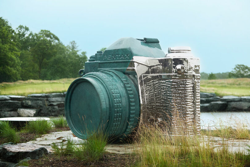 A large, metallic sculpture of a vintage Pentax camera is displayed outdoors amidst greenery and a body of water.