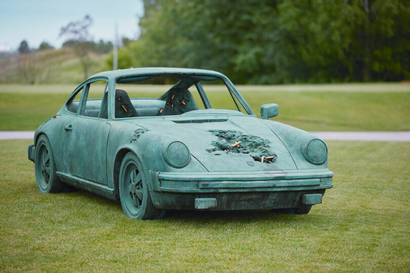 A bronze sculpture of a damaged Porsche car is placed on a grassy area, with noticeable crumples and holes in the body of the vehicle. Trees and a field are visible in the background.