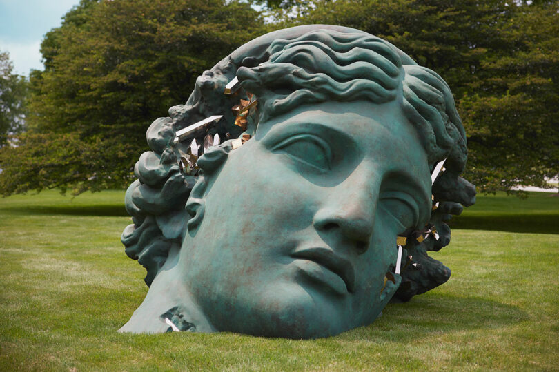 A large bronze head sculpture lies on a grassy field, with shards protruding from one side. Trees are visible in the background.